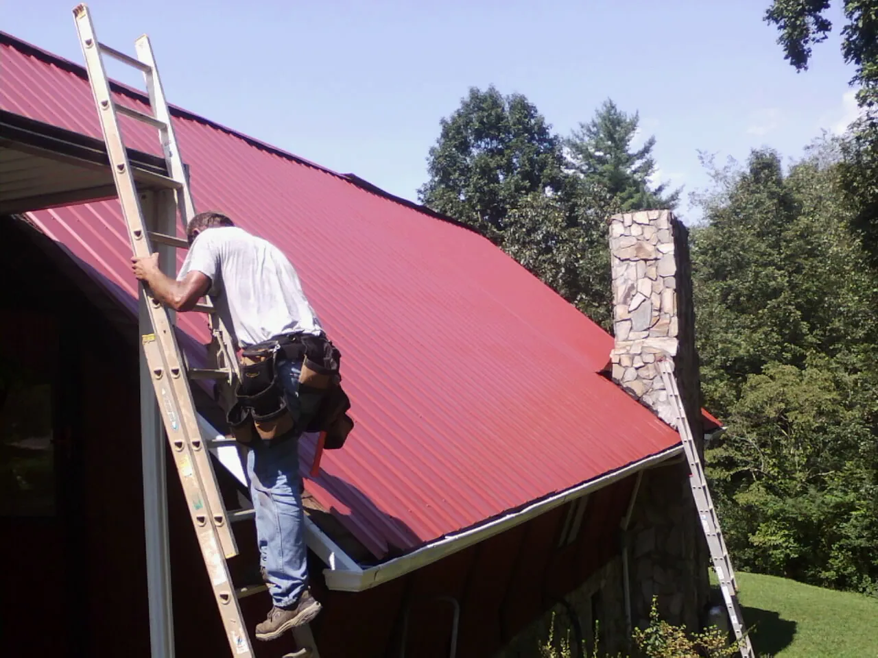 Roofer on a ladder beside a red metal roof and stone chimney — Soffit & Fascia Repair by Roofing Brothers in Lavalette